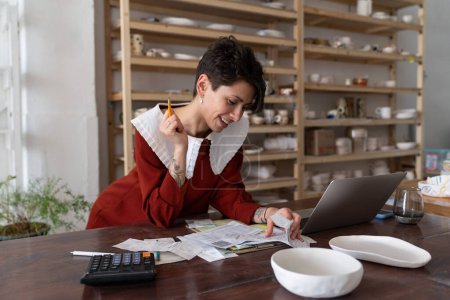 Bookkeeping in small business. Satisfied successful businesswoman ceramic studio owner calculating revenue, paying bills or taxes online, analyzing material costs to produce pottery. Entrepreneurship