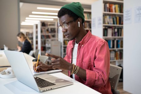 Photo for Concentrated focused African American student man in headphones look at laptop screen connection, discuss lecture educational material or studies foreign language with tutor teacher using video call. - Royalty Free Image
