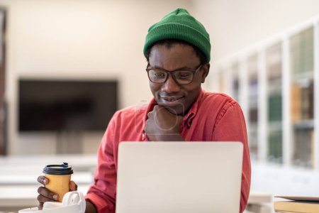 Téléchargez les photos : Pensif homme afro-américain avec café en tasse de papier se sentant satisfait des résultats du test, fier du travail de projet fait, tenir menton détendu. Réfléchir souriant mec noir pigiste regarde l'écran d'ordinateur portable. - en image libre de droit