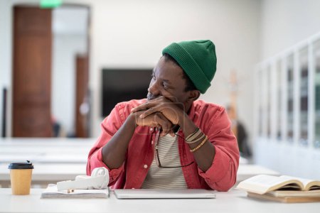 Foto de El afro-americano estresado se ve devastado mientras se sienta en la biblioteca universitaria y piensa en un posible fracaso en el examen. Hombre estudiante cansado frustrado por la falta de beca odia el sistema educativo - Imagen libre de derechos