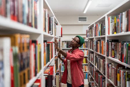 Photo for Thoughtful Black student guy searching materials for educational research in college library. Young African man choosing book for reading in bookstore, selective focus. Literature and education - Royalty Free Image