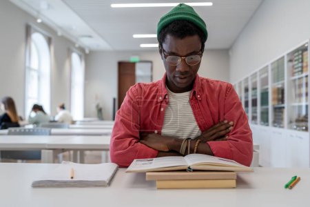 Photo for Book focused African American student guy reads study information with arms crossed in preparation for difficult exam in university library. Thoughtful man in eyeglasses reads textbook difficult task - Royalty Free Image
