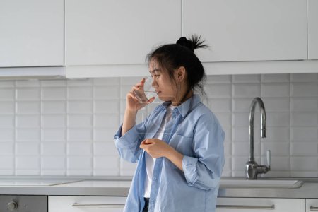 Photo for Sick unhealthy young Asian woman drinking water to to prevent fluid loss or dehydration during flu, depressed Korean girl standing in kitchen at home taking antidepressants, coping with depression - Royalty Free Image