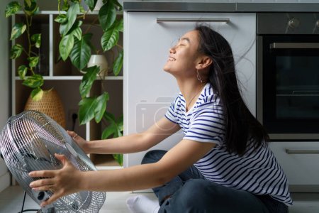Foto de Feliz alegre joven asiática sentada frente al ventilador de piso eléctrico en casa, enfriándose, refrescante durante los días calurosos de verano en apartamento sin aire acondicionado en verano - Imagen libre de derechos