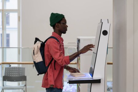 Photo for Concentrated African American man student stands near electronic terminal to check class timetable in college. Young attentive guy with backpack looks at monitor for self-service machines in library - Royalty Free Image