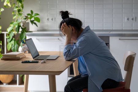 Photo for Tired devastated Asian woman bowing head wearily sits at desk with laptop thinks about project deadline about difficult work tasks. Overworked female frustrated by lack of money debts mortgage at home - Royalty Free Image