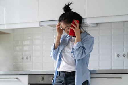 Photo for Sad depressed young Asian woman standing at home talking phone, hearing bad news, calling psychologist to receive mental health support, frustrated female making phone call, needs someone to talk to. - Royalty Free Image