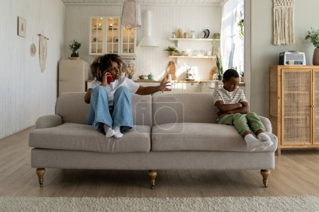 Photo for Annoyed angry African American mother sitting with offended upset son on sofa at home, talking on phone, discussing child behavior with spouse. Mom and kid conflict. Parents, children and discipline - Royalty Free Image