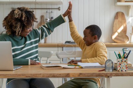 Joyful friendly mother and biracial son giving high five sitting at kitchen table celebrating success in study. Effective good cooperation result teamwork learn motivation in homeschooling with parent
