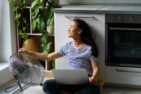 Arbeiten Sie bei Hitze von zu Hause aus. Glücklich lächelnde asiatische Remote-Arbeiterin arbeitet online am Laptop, sitzt auf dem Boden in der Nähe eines elektrischen Ventilators zu Hause, genießt die frische Luft, hält das Home Office in der Sommerhitze kühl