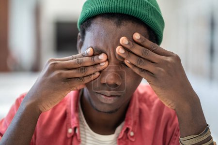 Téléchargez les photos : Homme afro-américain stressé souffrant de maux de tête ou de fatigue chronique au travail. Le noir épuisé tient la tête, ferme les yeux et s'énerve à cause des ennuis. Longue attente douloureuse, épuisement émotionnel. - en image libre de droit