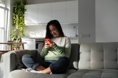 Photo for Carefree Asian woman with phone in hands sits on sofa enjoying visiting social networks. Cheerful young Japanese girl digitally addicted to gadgets spending all free time surfing internet - Royalty Free Image
