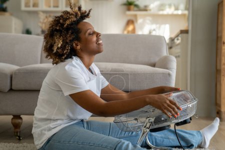Téléchargez les photos : Overheating in homes. Happy young African American woman sits in front of electric fan on floor in living room, enjoying blowing cool air, overheated female cooling down at home after being in heat - en image libre de droit