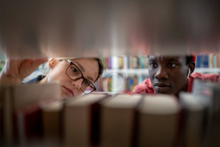 Photo for Two diverse students African guy and Caucasian girl studying together in library, standing between bookshelves and searching sources for research project. Collaborative learning in higher education - Royalty Free Image