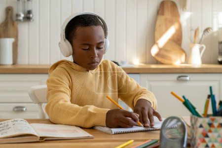 Téléchargez les photos : Focalisé impliqué enfant afro-américain auto-étude écrire avec manuel, tutoriel à la table de cuisine seule maison. Les éducations concentrées écolier noir dans les écouteurs préparent les devoirs de test scolaire - en image libre de droit