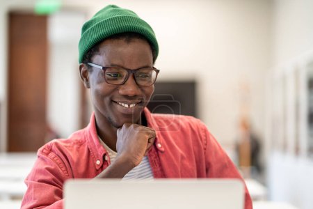Téléchargez les photos : Joyeux étudiant africain heureux gars regardant l'écran d'ordinateur portable tout en étudiant à la bibliothèque de l'université, foyer sélectif. Positif jeune homme noir profiter e-learning, regarder webinaire intéressant sur ordinateur - en image libre de droit
