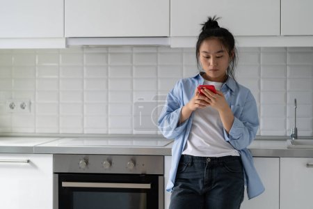 Photo for Surfing social media addicted Asian Korean woman stand at home puts things off until later, not concentrate and look away from mobile phone. Chinese girl use mobile phone in kitchen read online recipe - Royalty Free Image