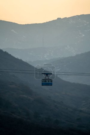 Photo for Public transport for the highlands. Blue old cabin of cable car moves against backdrop of crested mountains in fog.Climb to top hill with funicular. Misty place pastels colour with strung wires in air - Royalty Free Image