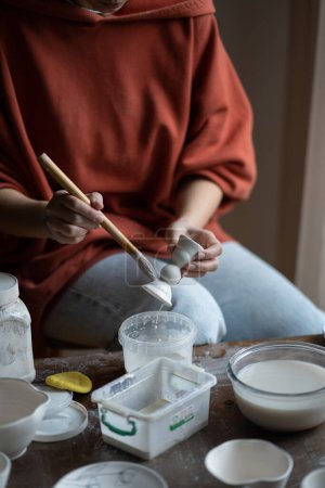 Foto de Female ceramicist or potter holding paintbrush applying glaze on ceramic pieces while sitting at work table in studio, cropped photo. Production of handmade tableware. Pottery therapy in mental health - Imagen libre de derechos