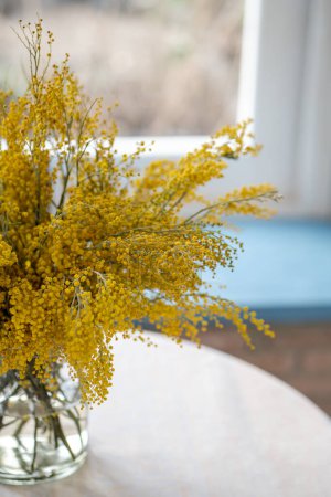 Téléchargez les photos : Bouquet de fleurs de Mimosa dans un vase en verre sur table à la maison, mise au point douce. Bouquet de printemps avec Acacia dealbata, faible profondeur de champ. 8 mars, concept de Pâques. - en image libre de droit