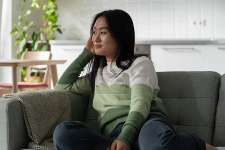 Foto de Positiva mujer asiática alegre se relaja se sienta en el sofá disfrutando del silencio y la soledad. Feliz niña coreana despreocupada zoomer sonrisas apoyándose de cabeza en la mano soñando con futuras vacaciones o vacaciones - Imagen libre de derechos