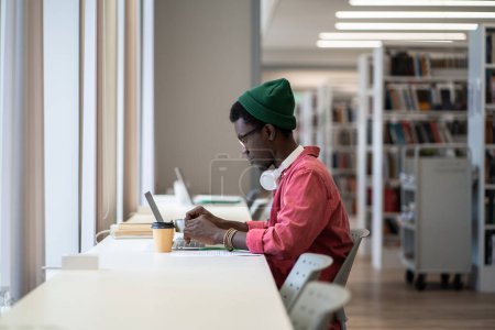 Téléchargez les photos : Focalisé noir assis au bureau avec ordinateur portable en utilisant la base de données de la bibliothèque en ligne, vue latérale. Étudiant afro-américain de sexe masculin écrivant des cours, se préparant aux examens universitaires. Internet dans l'éducation concept - en image libre de droit