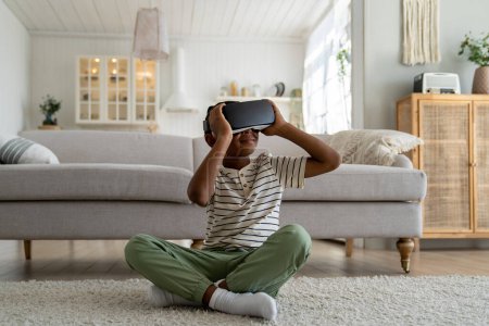 Photo for Pleased smiling African American schoolboy measuring metaverse immersing in virtual world touch goggles try to explore new 3D reality glasses. Carefree little boy online study in VR headset at home - Royalty Free Image