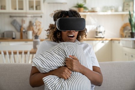 Foto de Mujer afroamericana asustada con auriculares de realidad virtual se sienta en el sofá de casa sintiéndose emocionada, abrumada por ver películas de terror. Afectado hembra joven negro en gafas 3D sostienen almohada del juego temido - Imagen libre de derechos