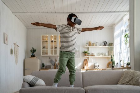 Photo for Carefree smiling African American child boy play video game with virtual reality goggles pretending to be airplane with arms outstretched. Joyful kid son have fun in VR headset involved in metaverse - Royalty Free Image