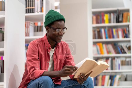 Photo for Interested African American student guy reading book in university library. Lover of literature spends time reading an interesting novel in bookstore. Black man in glasses with book studying for exam. - Royalty Free Image