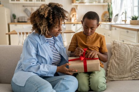 Photo for Caring loving African American mom greeting surprised small boy with present gift. Kind black woman give on birthday celebrate wrapped box with red ribbon to anticipating son sitting on couch at home - Royalty Free Image