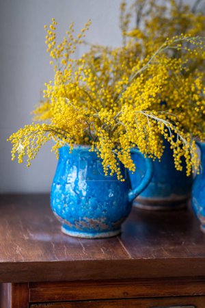 Foto de Ramillete de flores de Mimosa en jarrón de cerámica vintage azul sobre pared blanca en casa. Manojo de primavera con Acacia dealbata en superficie de madera, enfoque suave. 8 de marzo, concepto de Pascua - Imagen libre de derechos