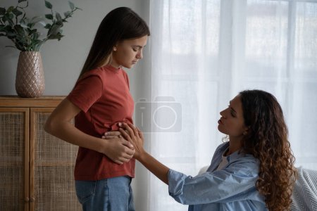 Photo for Depressed unhappy teenage girl near mom holding stomach complaining of pain after eating fast food. Kind caring woman nanny sits in house and looks with sympathy at child suffering from gastritis - Royalty Free Image