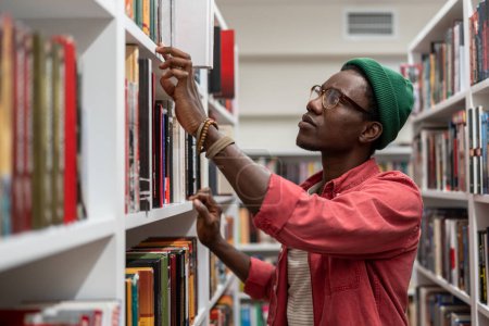 Photo for Interested Black student man choosing book for reading in bookstore. Focused young African American guy searching materials for educational research in college library. Literature and education - Royalty Free Image