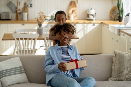 Photo for Loving mixed race small boy greet surprised mom with Mothers Day gives gift by closing eyes with hands. Smiling African American woman hold wrapped box with red ribbon congratulation from child son - Royalty Free Image