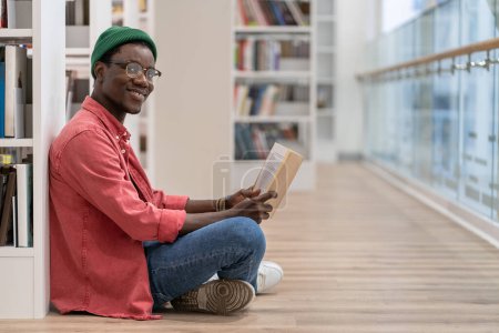 Photo for Black student man library visitor sitting on floor near bookcase with book in hands, looking at camera and smiling, African American guy literature lover in public library interior. Reading hobby - Royalty Free Image