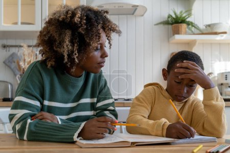 Téléchargez les photos : Enfant garçon biracial épuisé regarde livre d'étude tout en écoutant tuteur afro-américain femme à la leçon privée à domicile. Travail éducatif pour enfant concentré fils avec maman aider à la tâche de devoirs - en image libre de droit