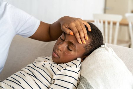 Photo for Sick African American child boy lies with closed eyes on couch feel unwell weakness. Closeup hand woman mom touch forehead check temperature unhealthy small son with fever symptom weariness. Childcare - Royalty Free Image