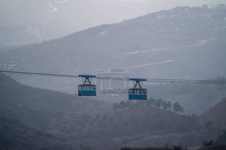 Photo for Two blue old cabins cable car moves against backdrop of crested mountains in fog. Climb to top hill with funicular. Misty beautiful place with strung wires in air. Public transport for the highlands. - Royalty Free Image