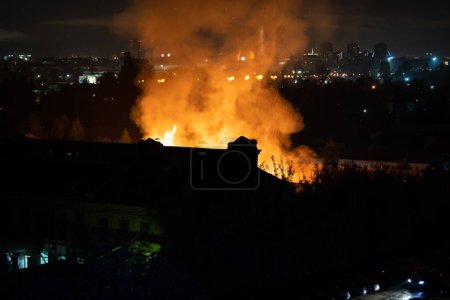 Edificio en llamas por la noche en la ciudad. Las llamas anaranjadas y el humo pesado saliendo de la casa dañada en llamas durante la noche. Peligro de incendio en los edificios concepto