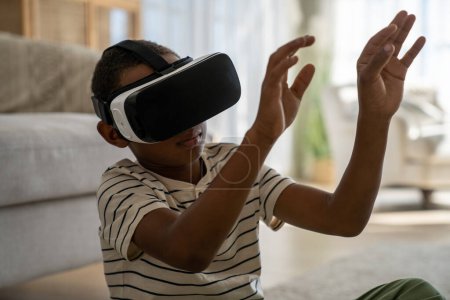 Foto de El niño afroamericano concentrado que mide la metáfora sumergiéndose en el aire táctil del mundo virtual con las manos intenta explorar nuevas gafas de realidad 3D. Pequeño colegial estudio en línea en VR auriculares en casa - Imagen libre de derechos