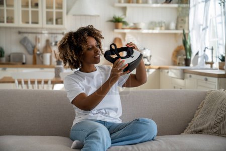 Foto de Experiencia VR. Curiosa mujer africana joven emocionada sosteniendo gafas 3D modernas mientras se sienta en el sofá en la sala de estar, preparándose para usar auriculares de realidad virtual. Tecnología del concepto futuro - Imagen libre de derechos
