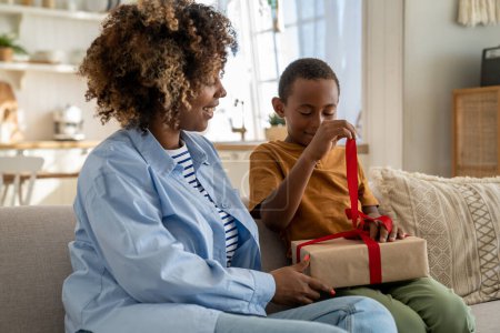 Téléchargez les photos : Aimer mère afro-américaine féliciter heureux petit fils avec cadeau d'anniversaire enveloppé avec ruban rouge. Célébrer ensemble maman noire et garçon enfant assis sur le canapé à la maison déballer boîte surprise - en image libre de droit