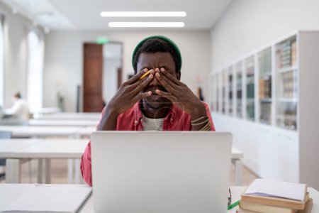 Photo for Exhausted African American student man sit at library desk with laptop feel unwell rub dry irritable eyes try concentrated. Tired black guy freelancer with strain tension migraine after computer work - Royalty Free Image