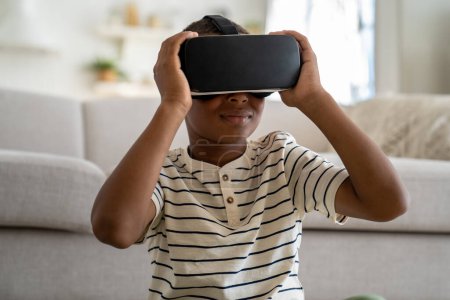 Photo for Curious African American kid boy wearing virtual reality 3D glasses online study simulation virtual world at home. Serious small schoolboy get experience in education knowledge with help VR goggles - Royalty Free Image