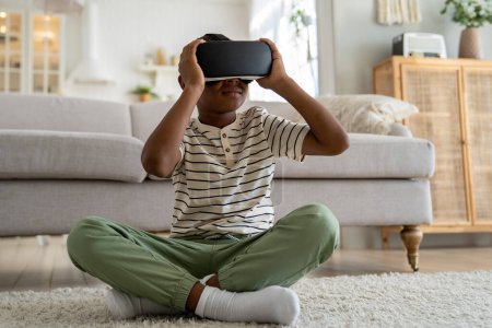 Photo for African American boy child sitting on floor in living room wearing virtual reality headset exploring virtual world. Schoolboy kid using VR technology for learning. Augmented reality for children - Royalty Free Image