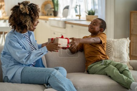 Photo for For the best mom ever. Proud happy African American boy son giving wrapped gift box to surprised excited mother, sitting together on couch at home. Child kid surprising mum on Mothers day - Royalty Free Image