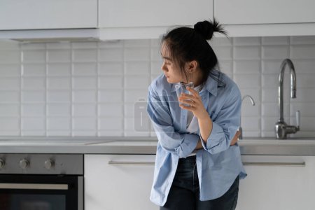 Foto de Mujer asiática deprimida infeliz que tiene problemas digestivos de pie en la cocina en casa con un vaso de agua. Sufrimiento señora china sostiene el estómago y se inclina debido al dolor en el tracto gastrointestinal - Imagen libre de derechos