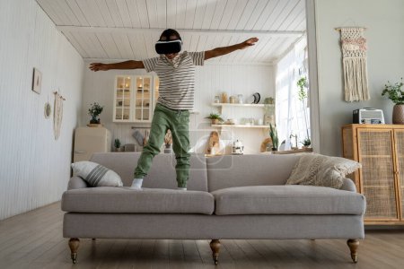 Foto de Niño africano despreocupado que utiliza casco VR para la diversión y el entretenimiento, niño feliz de pie en el sofá jugando inmersivo juego de vuelo virtual 3D, volando en el mundo metaverso. Niños en un ambiente inmersivo - Imagen libre de derechos