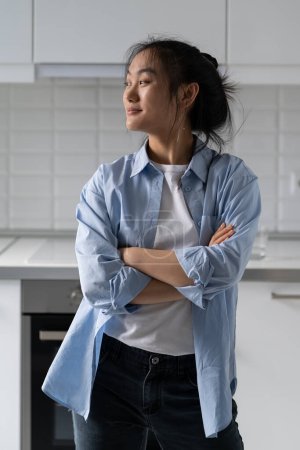 Foto de Relaxed dreamy Asian woman smiling and looking away thinking about future plans standing in kitchen. Optimistic young korean girl posing with arms crossed remembering happy moments from vacation - Imagen libre de derechos